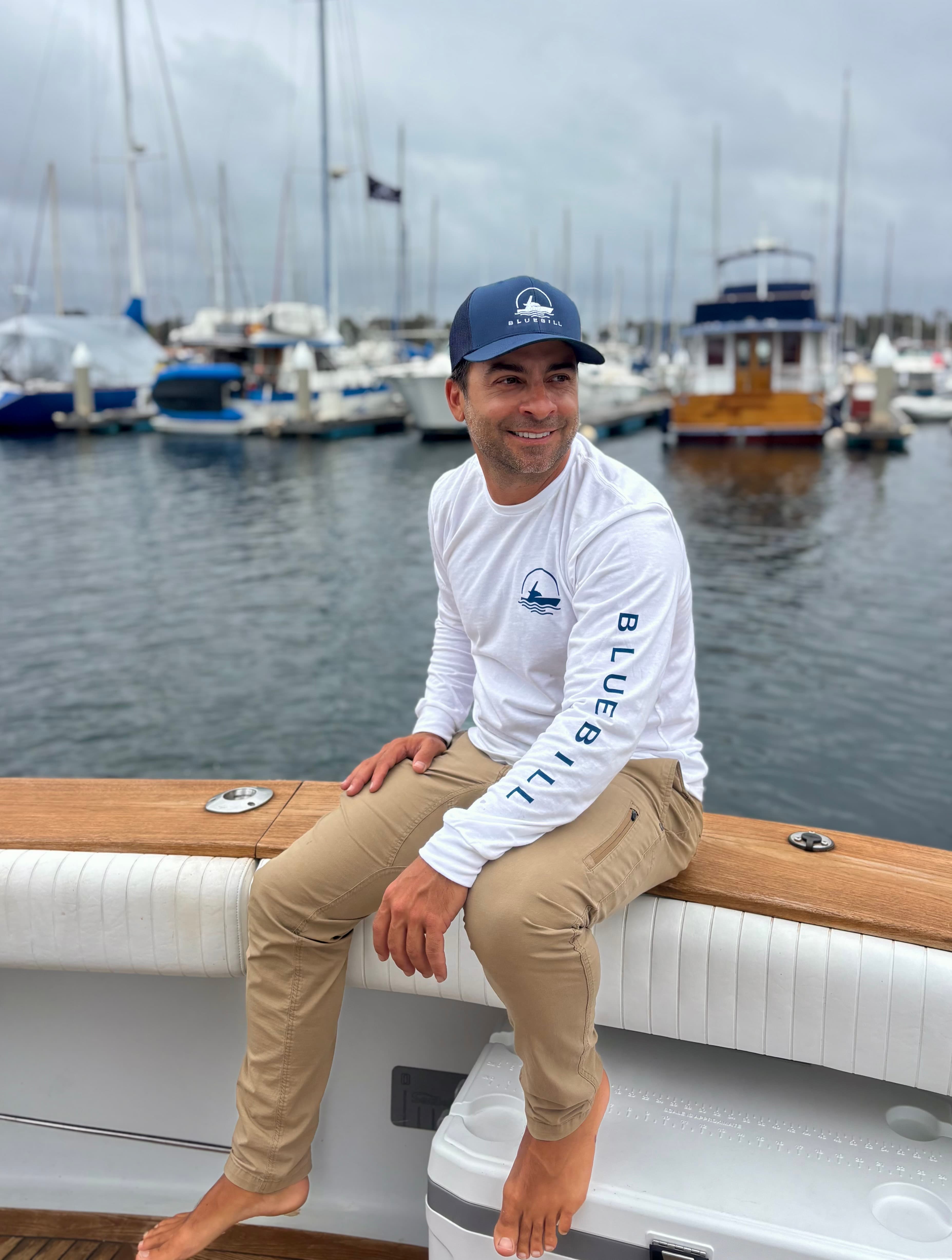 Man on a boat gunwale wearing a white Bluebill shirt and navy cap